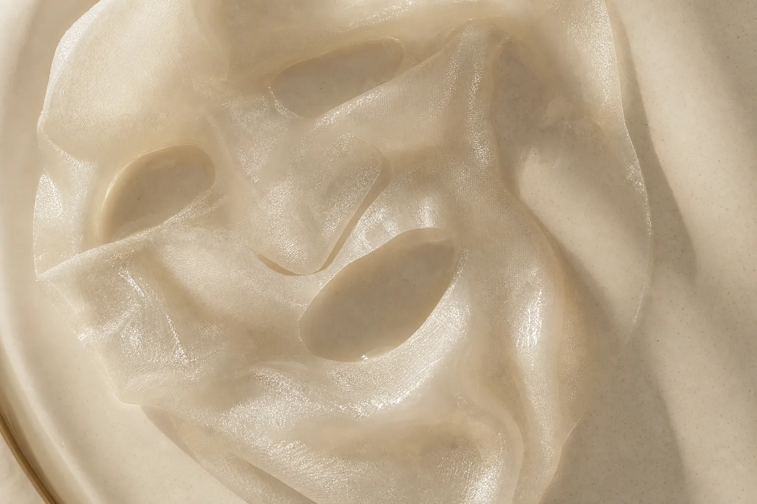 A translucent essence-soaked sheet mask fabric gently unfolded on a warm cream surface, editorial macro photography